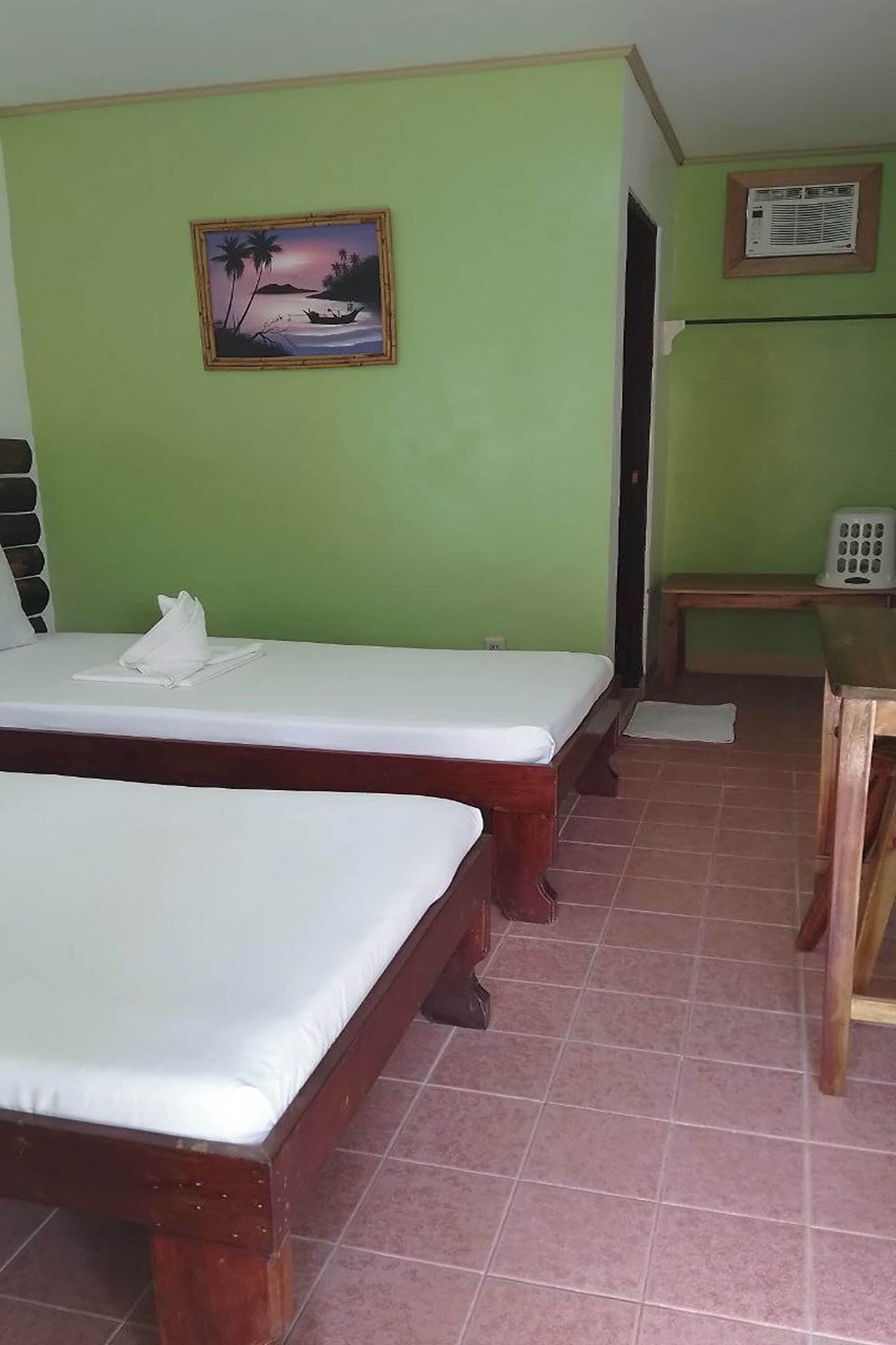 A room with two wooden beds, a green wall, a painting of a beach scene, and a small table with a chair.