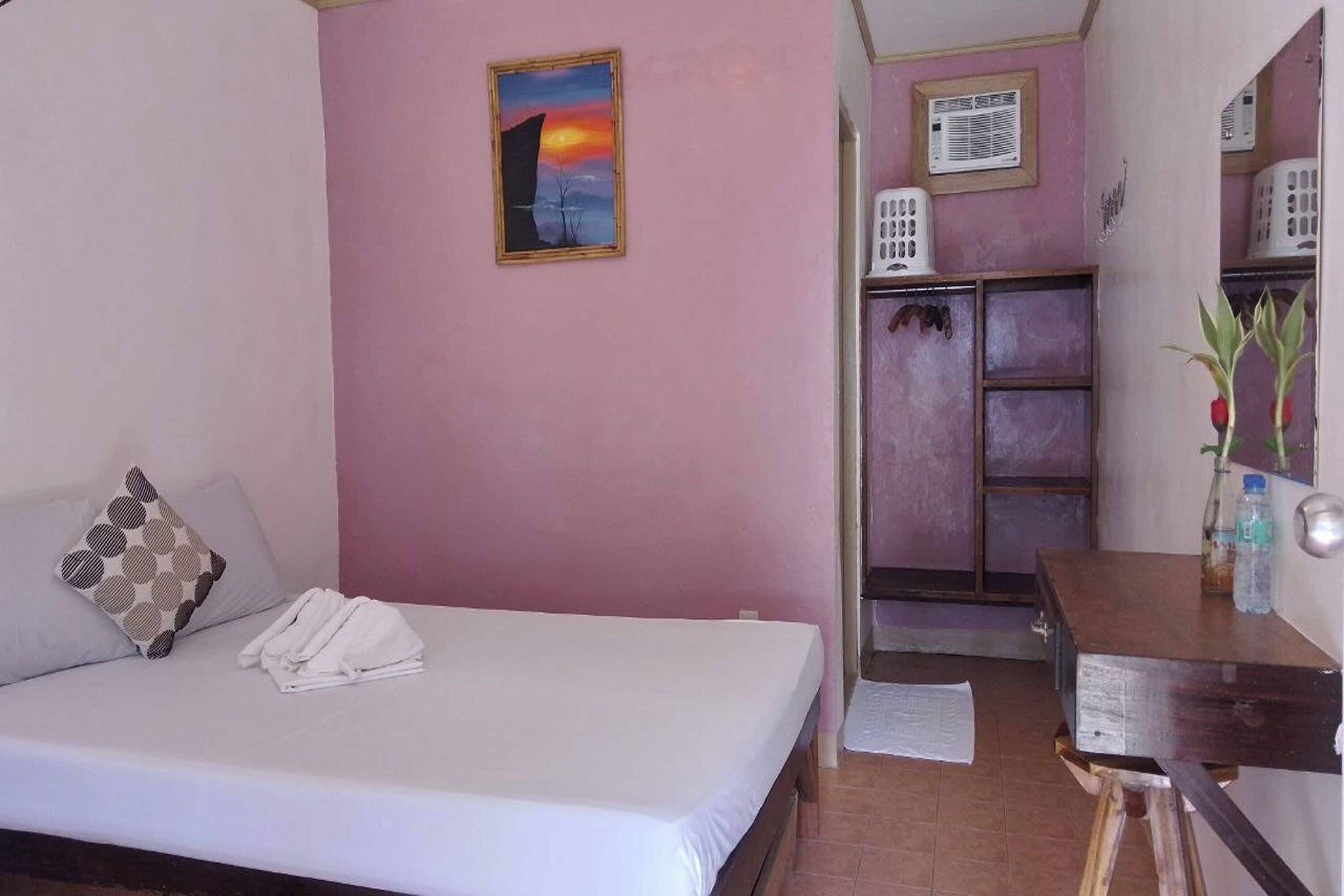 A bedroom with a bed, pillows, a painting, a desk with a lamp, and a bathroom with a shower and toilet.