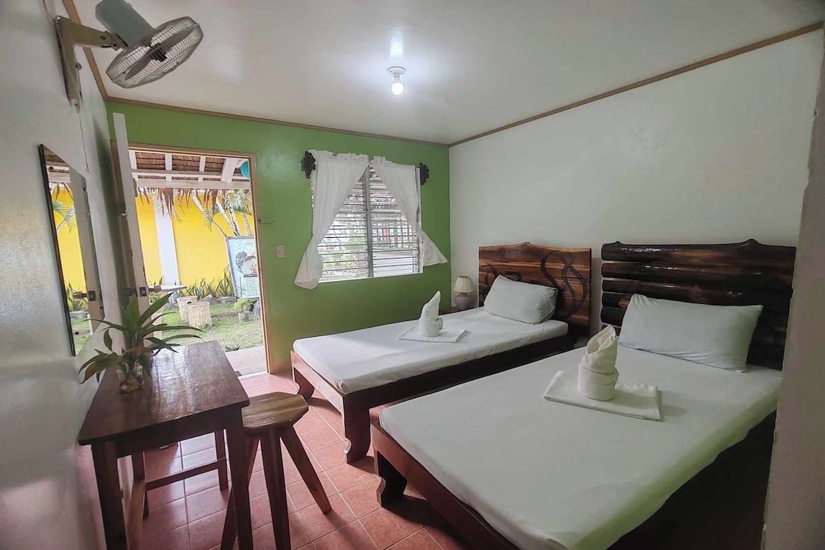 Una habitación con dos camas, una mesa, un ventilador y una ventana con cortinas.