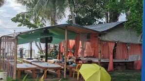 Un espacio de estar al aire libre de estilo rústico con mesas y bancos de madera, algunos cubiertos por un toldo verde y otros con tela naranja.