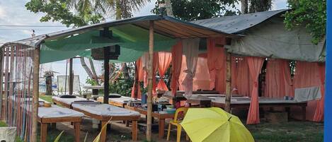 A rustic outdoor seating area with wooden tables and benches, some covered by a green canopy and others by orange fabric.