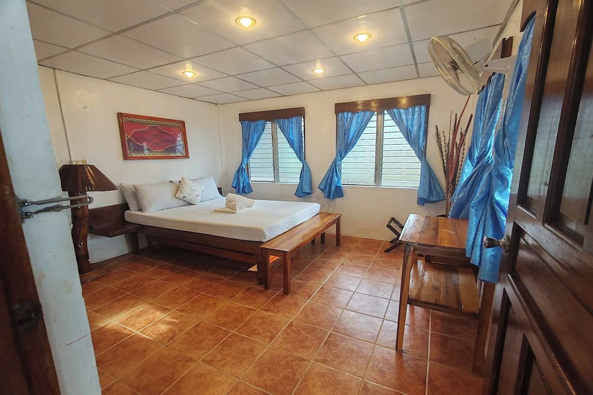 Un dormitorio con cama, un banco de madera, un escritorio y un ventilador.