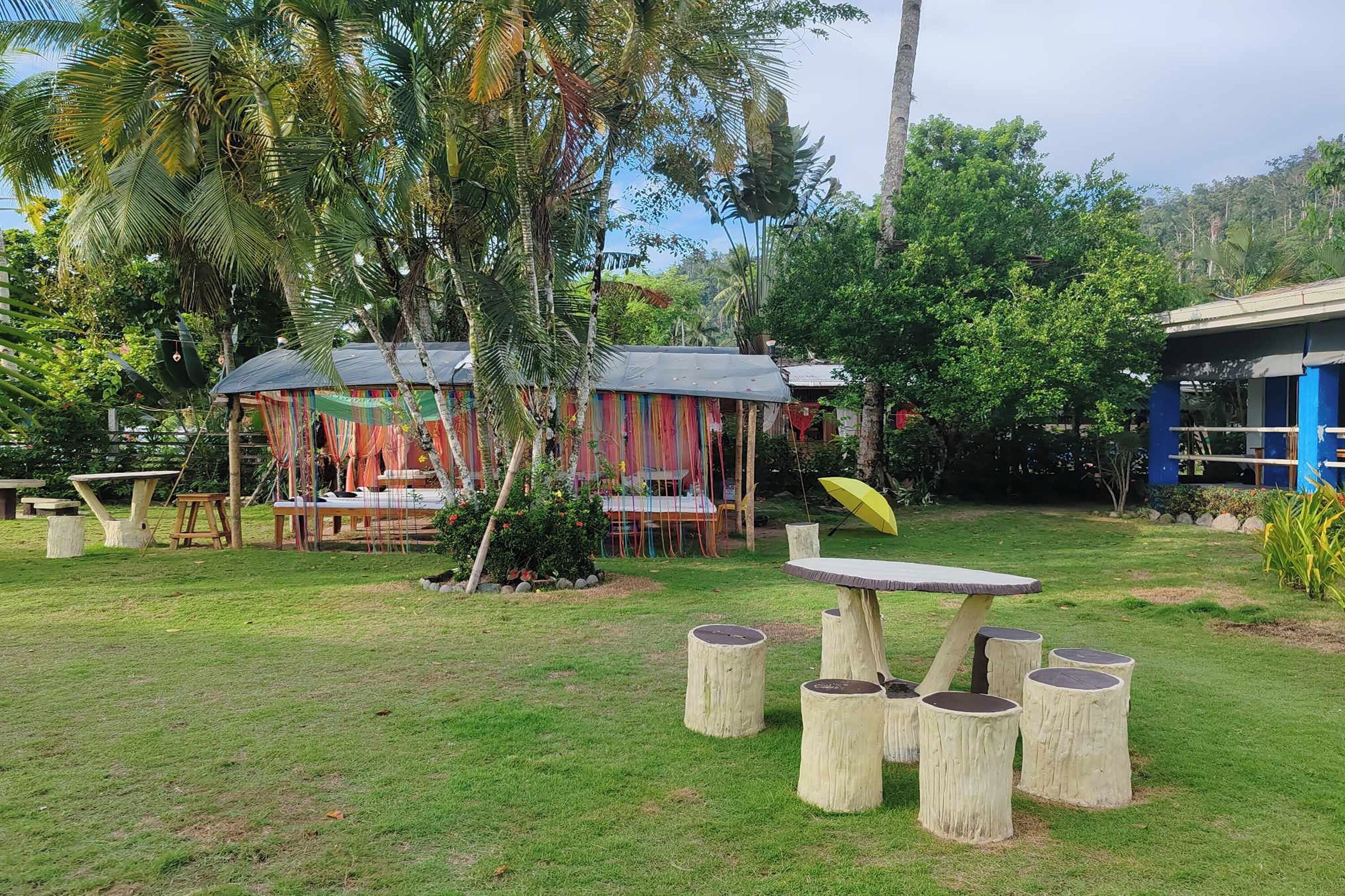 A tropical outdoor area with wooden tables and benches, surrounded by lush greenery and palm trees.