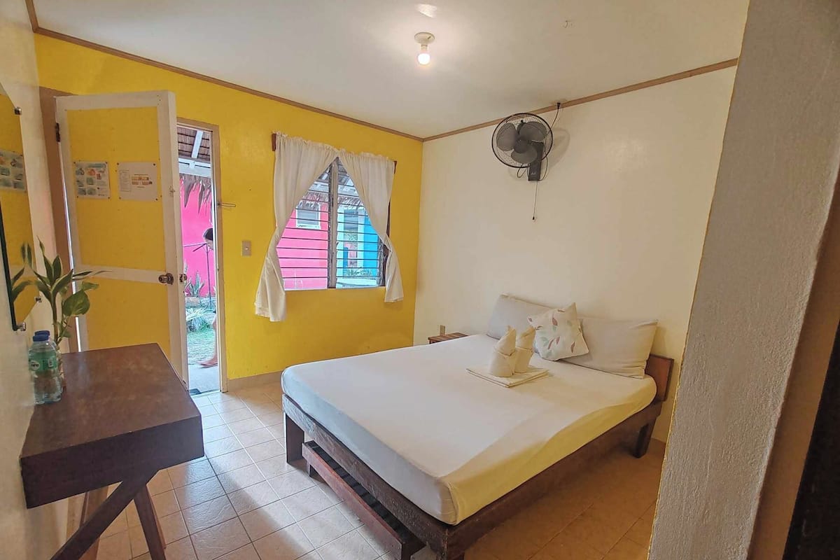 Una habitación con cama, escritorio, ventilador y ventana con cortinas.