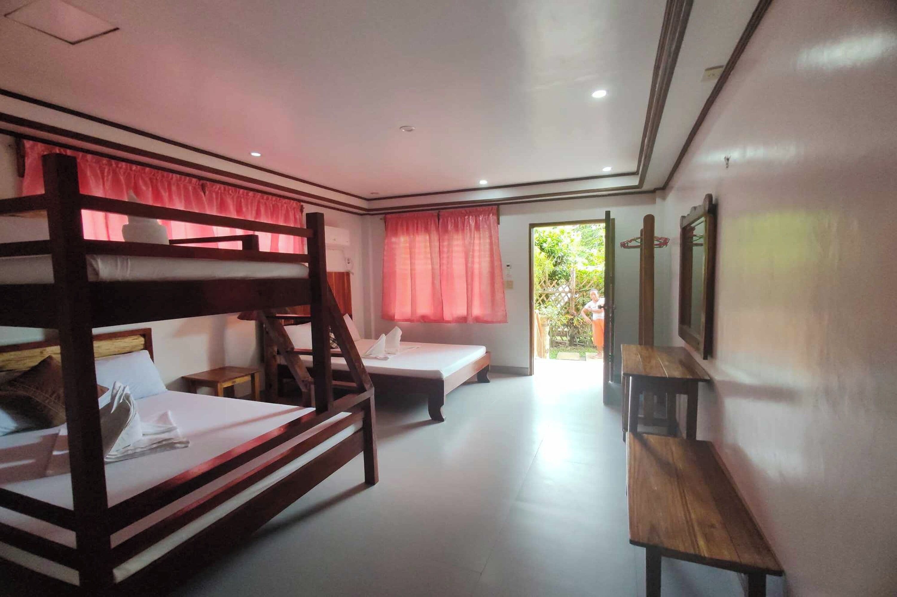 Habitación con literas, una cama individual, una mesita pequeña y un banco. Hay una ventana con cortinas y una puerta que da al exterior.