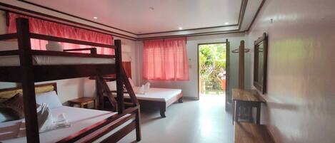 Habitación con literas, una cama individual, una mesita pequeña y un banco. Hay una ventana con cortinas y una puerta que da al exterior.