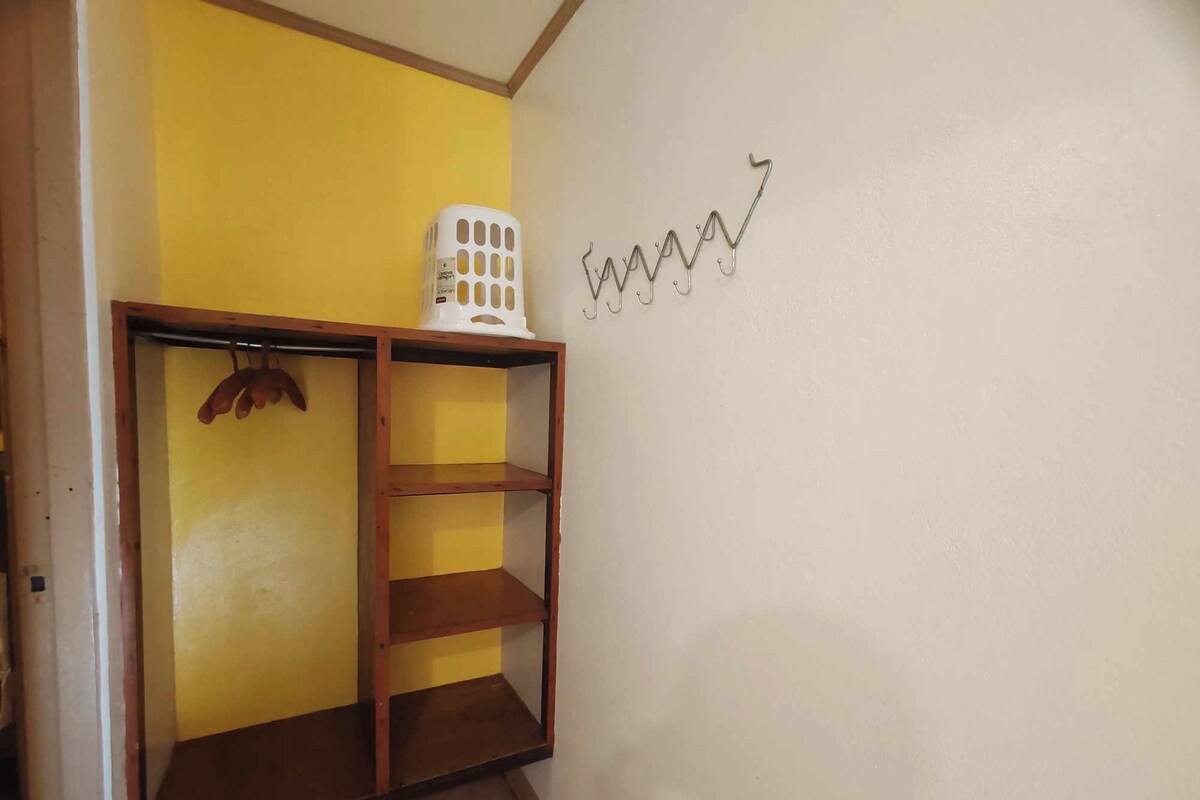 A room with a wooden shelf unit against a yellow wall, a white laundry basket on top, and a wall-mounted hook.
