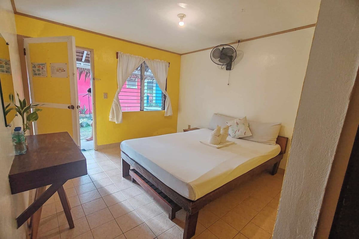 Una habitación con cama, escritorio, ventilador y ventana con cortinas.