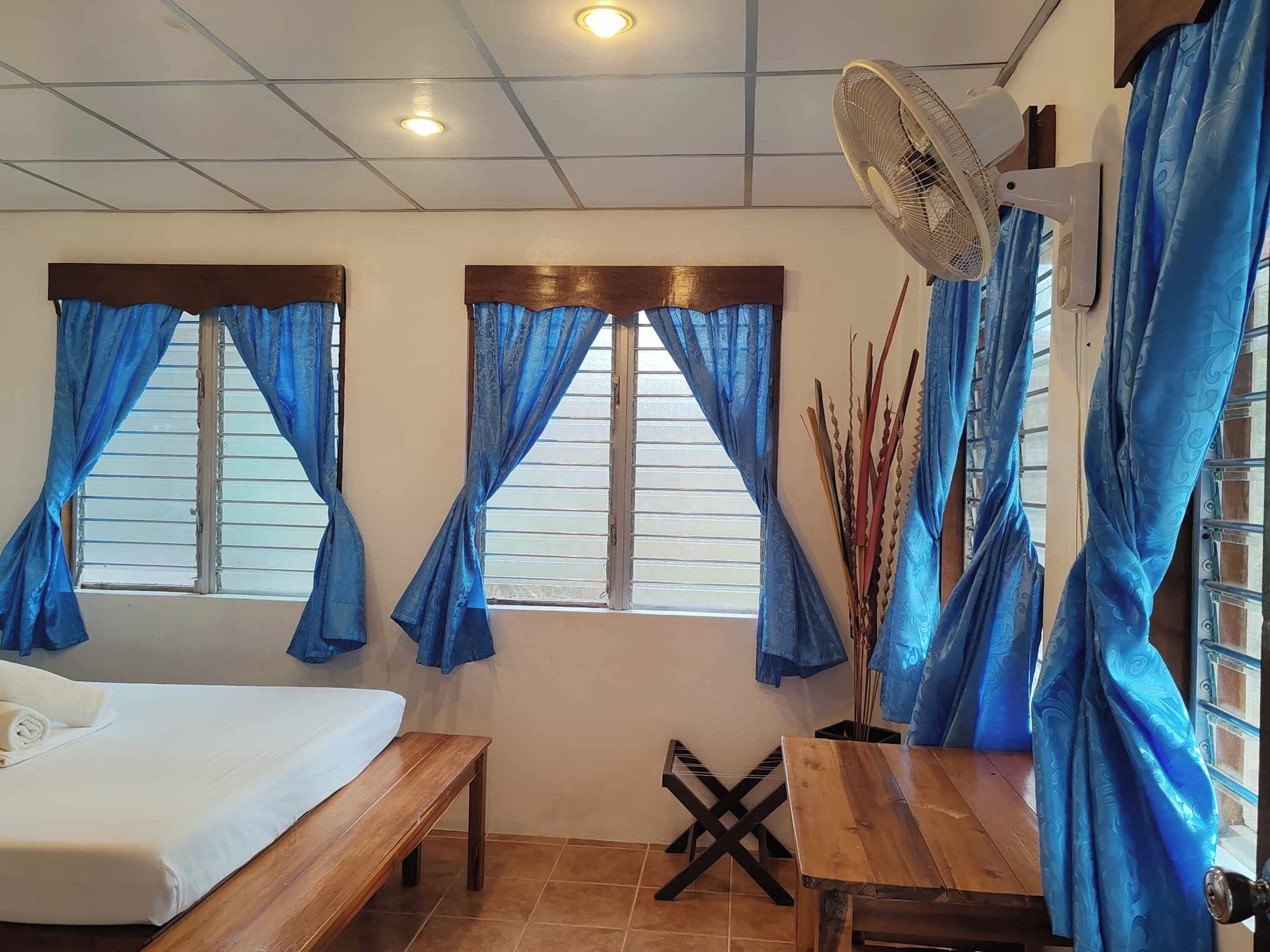 A room with a bed, a wooden table, a bench, a fan, and windows with blue curtains.