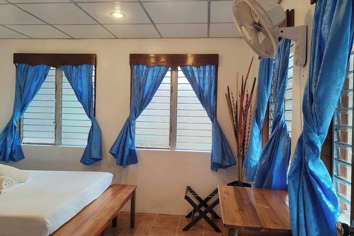 Una habitación con cama, una mesa de madera, un banco, un ventilador y ventanas con cortinas azules.