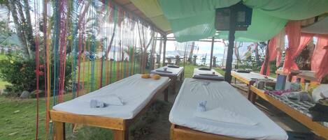 A covered outdoor relaxation area with wooden benches and white mattresses, surrounded by colorful fabric decorations and greenery.