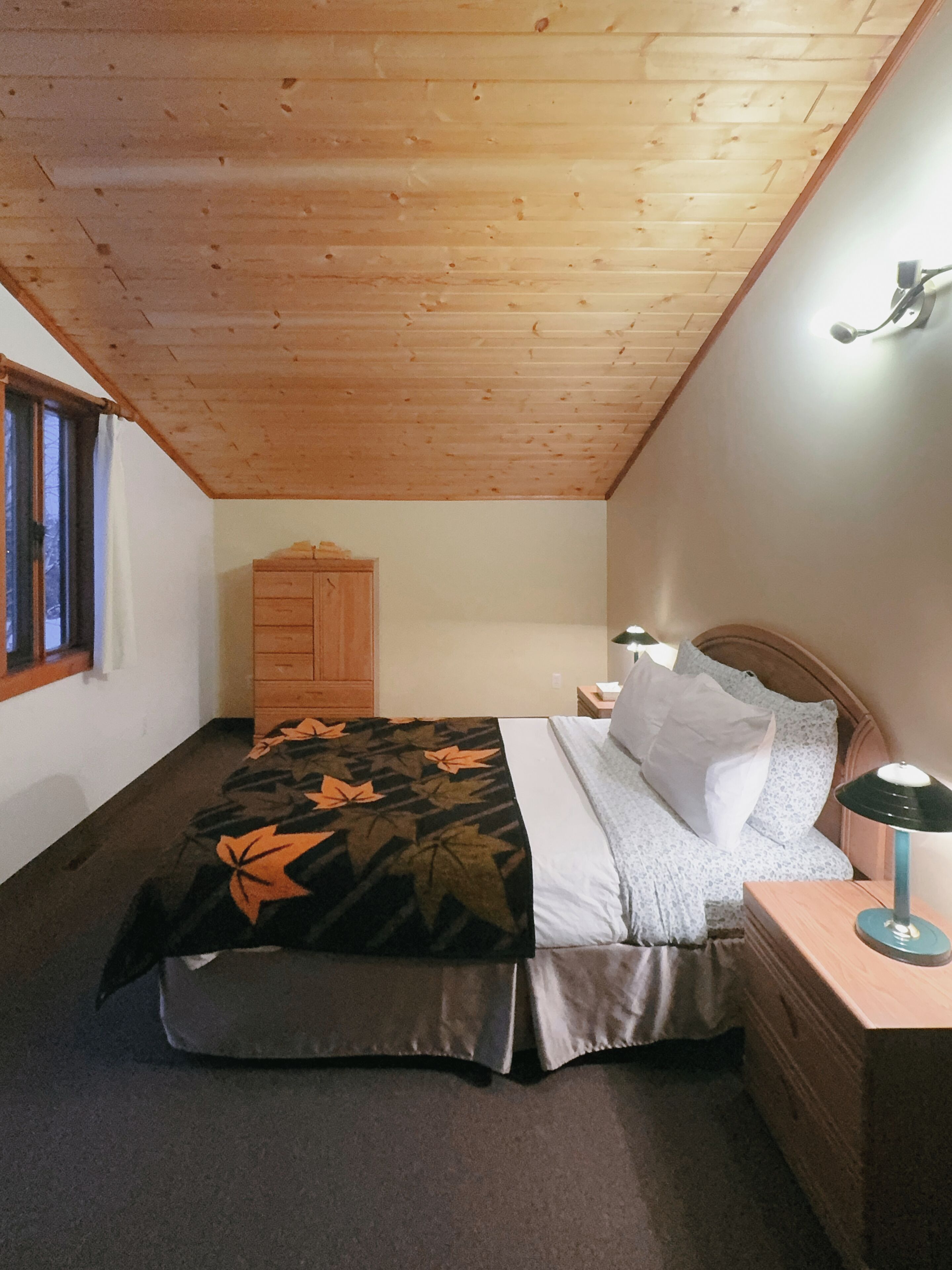 A bedroom with a wooden ceiling, a bed with a floral bedspread, a nightstand with a lamp, and a wooden wardrobe.