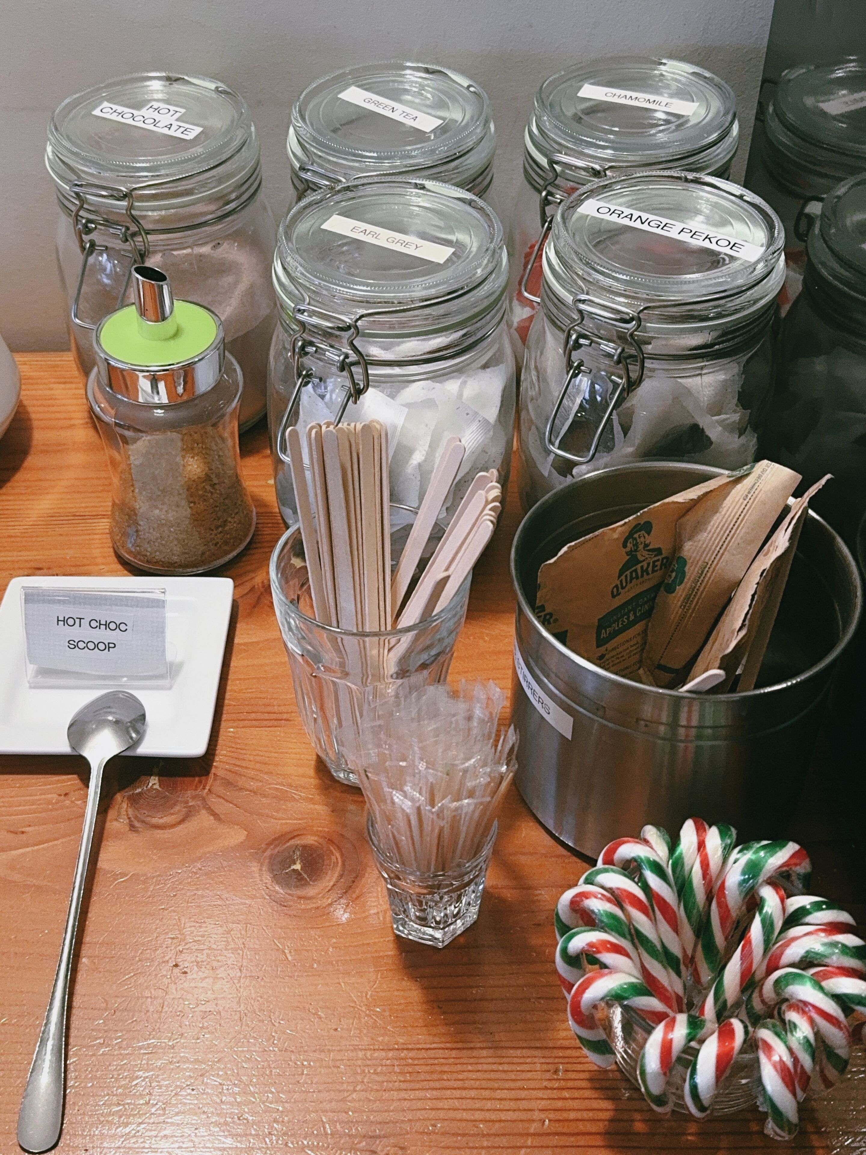 Potes de vidro com etiquetas, um recipiente de metal, uma colher e bastões de candy cane sobre uma superfície de madeira.