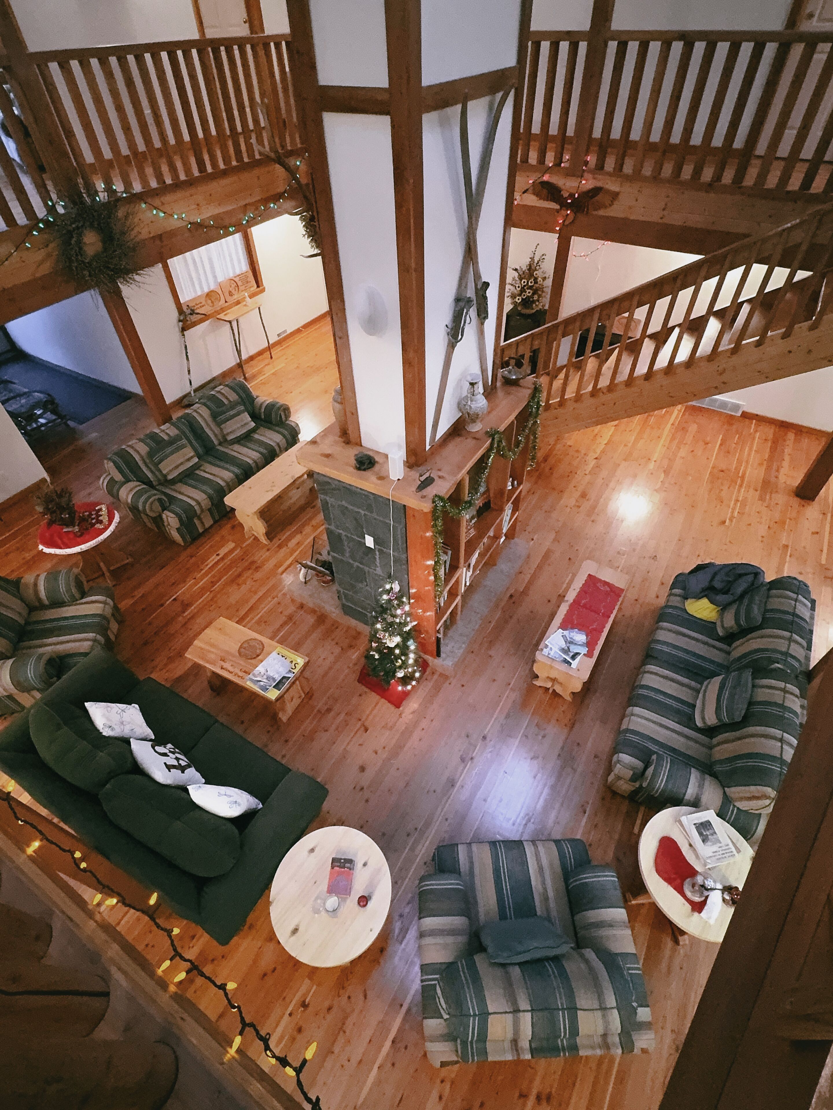 Un vaste espace de vie de style loft, doté de hauts plafonds, de poutres apparentes et d’une cheminée centrale. On y trouve plusieurs coins salon avec des canapés et une table basse. Un petit sapin de Noël apporte une touche festive.