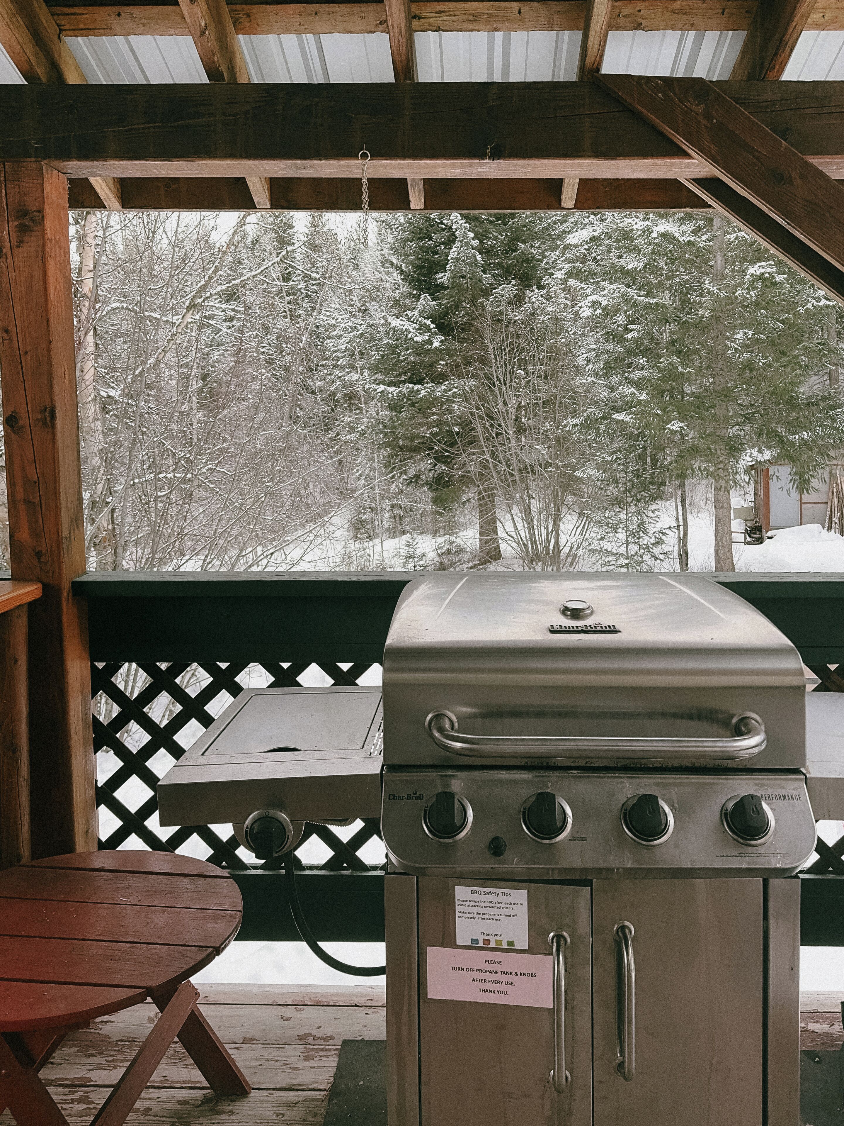 A covered outdoor grill area with a table and a view of snow-covered trees.