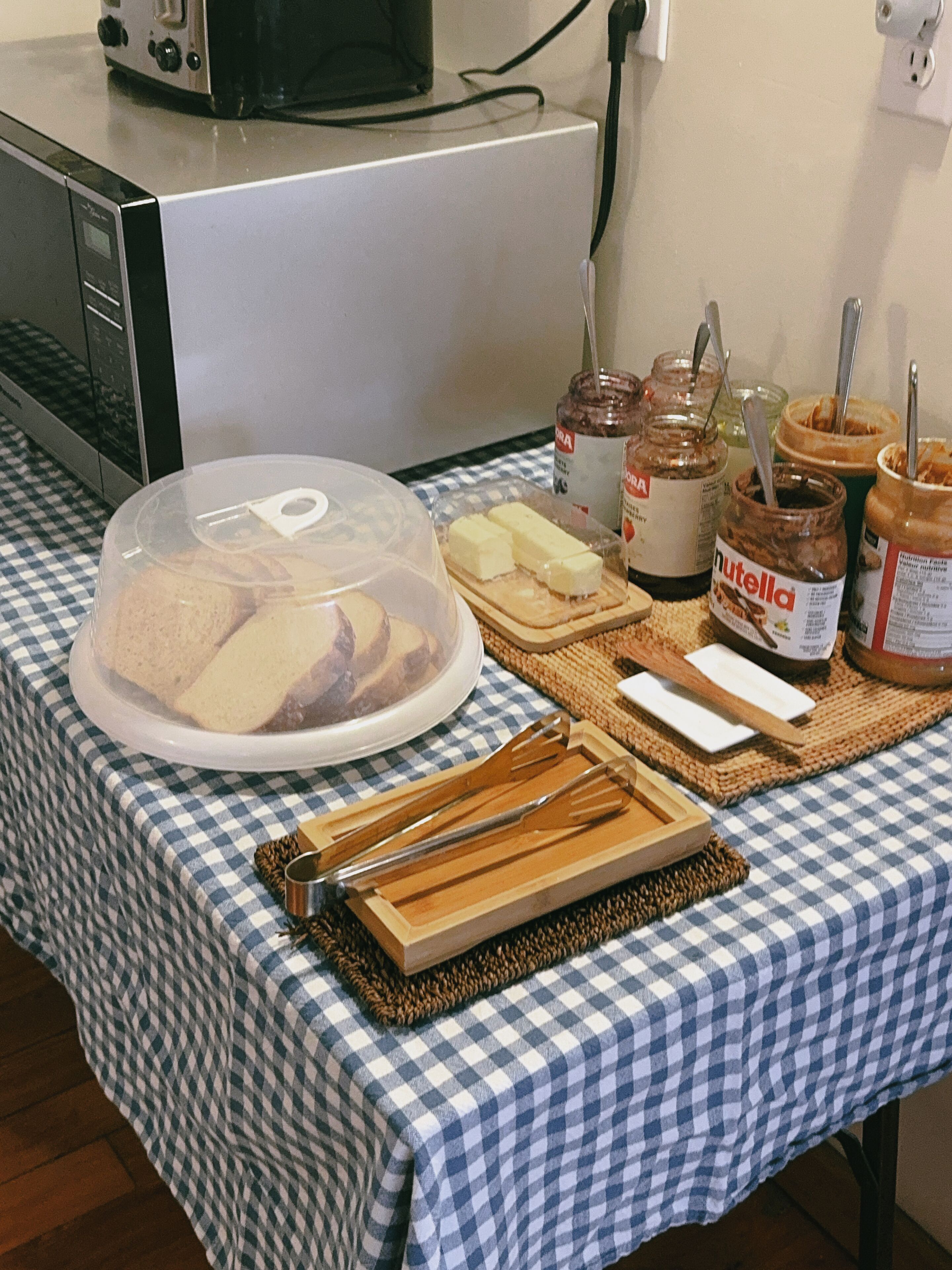 Une table recouverte d’une nappe à carreaux, un récipient hermétique contenant de la nourriture, du beurre, du Nutella et un couteau posés sur une planche en bois.