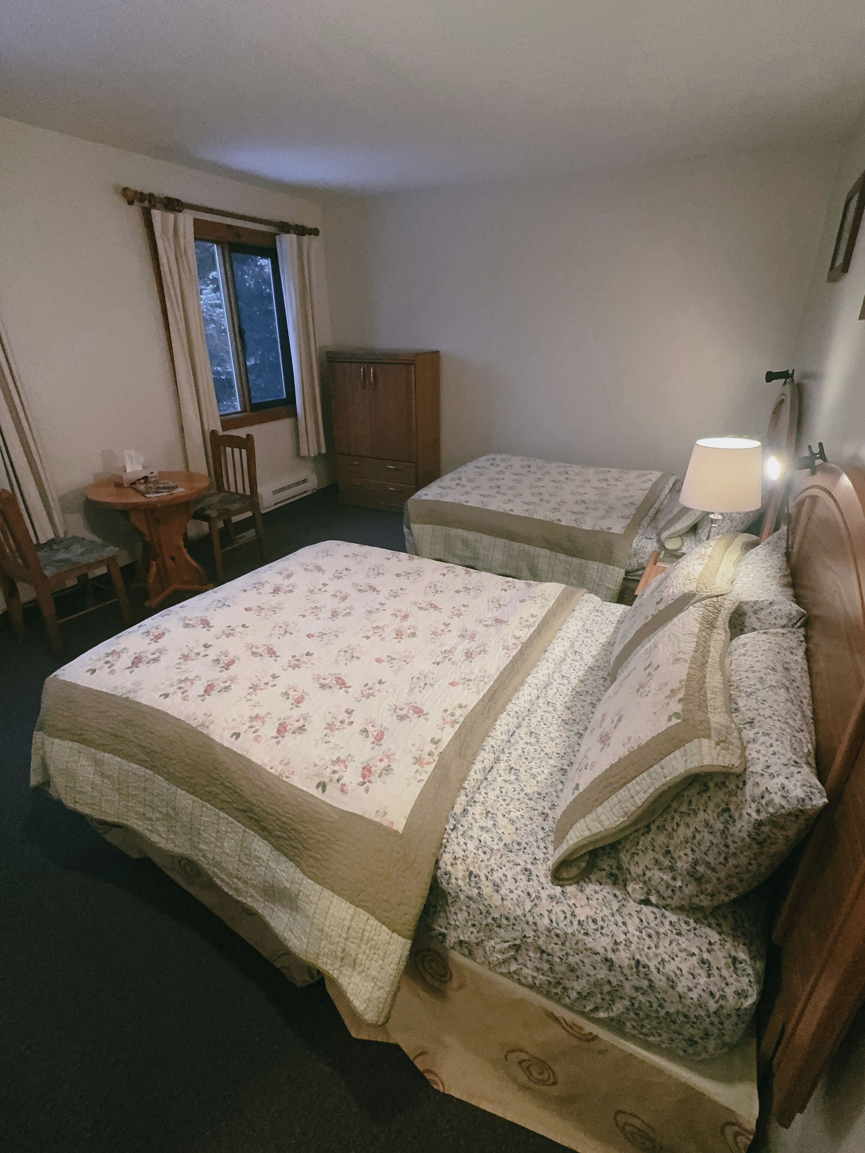Une chambre d’hôtel avec deux lits, une petite table, une chaise, une armoire et une fenêtre avec des rideaux.