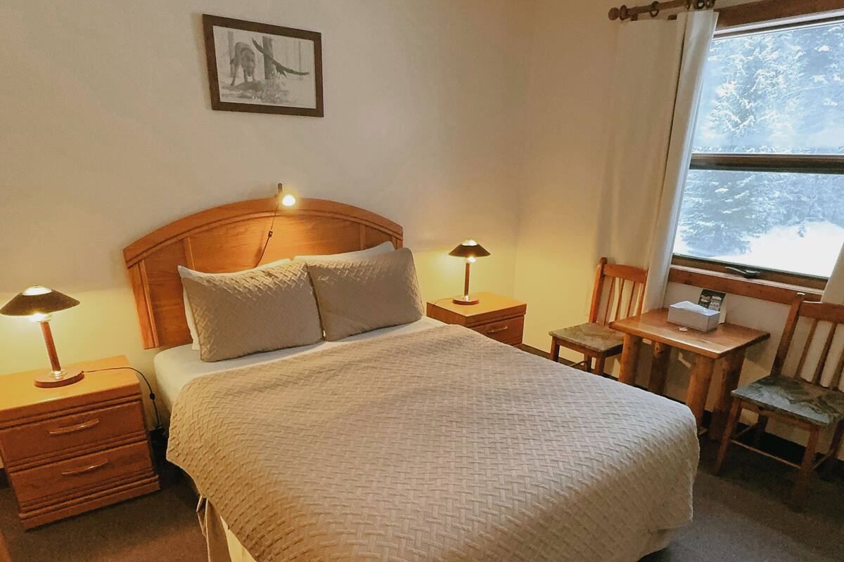 A neatly arranged bedroom with a large bed, two bedside tables with lamps, a small wooden table with a chair, a window with a view of trees, and a framed picture on the wall.