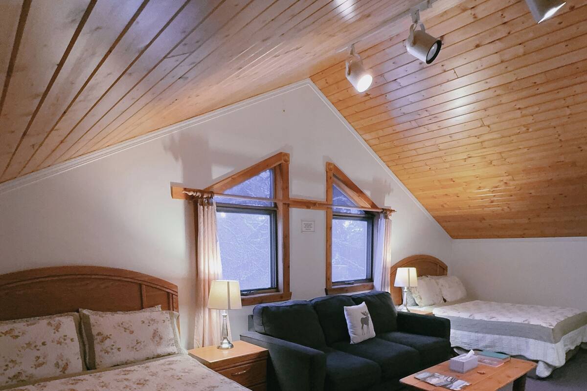 A cozy room with a bed, a sofa, and a small table. There are windows and a wooden ceiling.