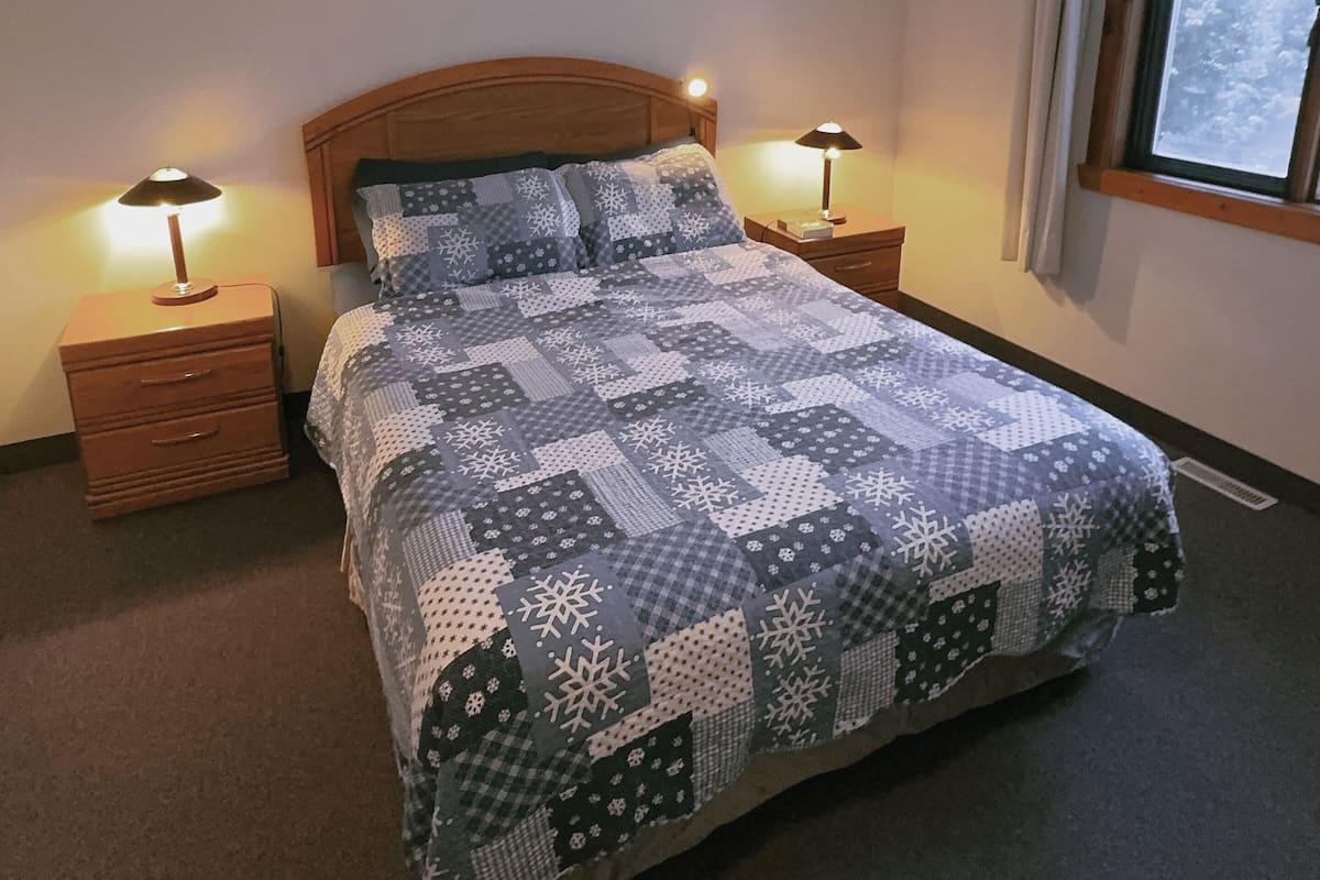 A bedroom with a wooden headboard, a quilted bedspread, two bedside tables with lamps, a framed picture on the wall, and a window with curtains.