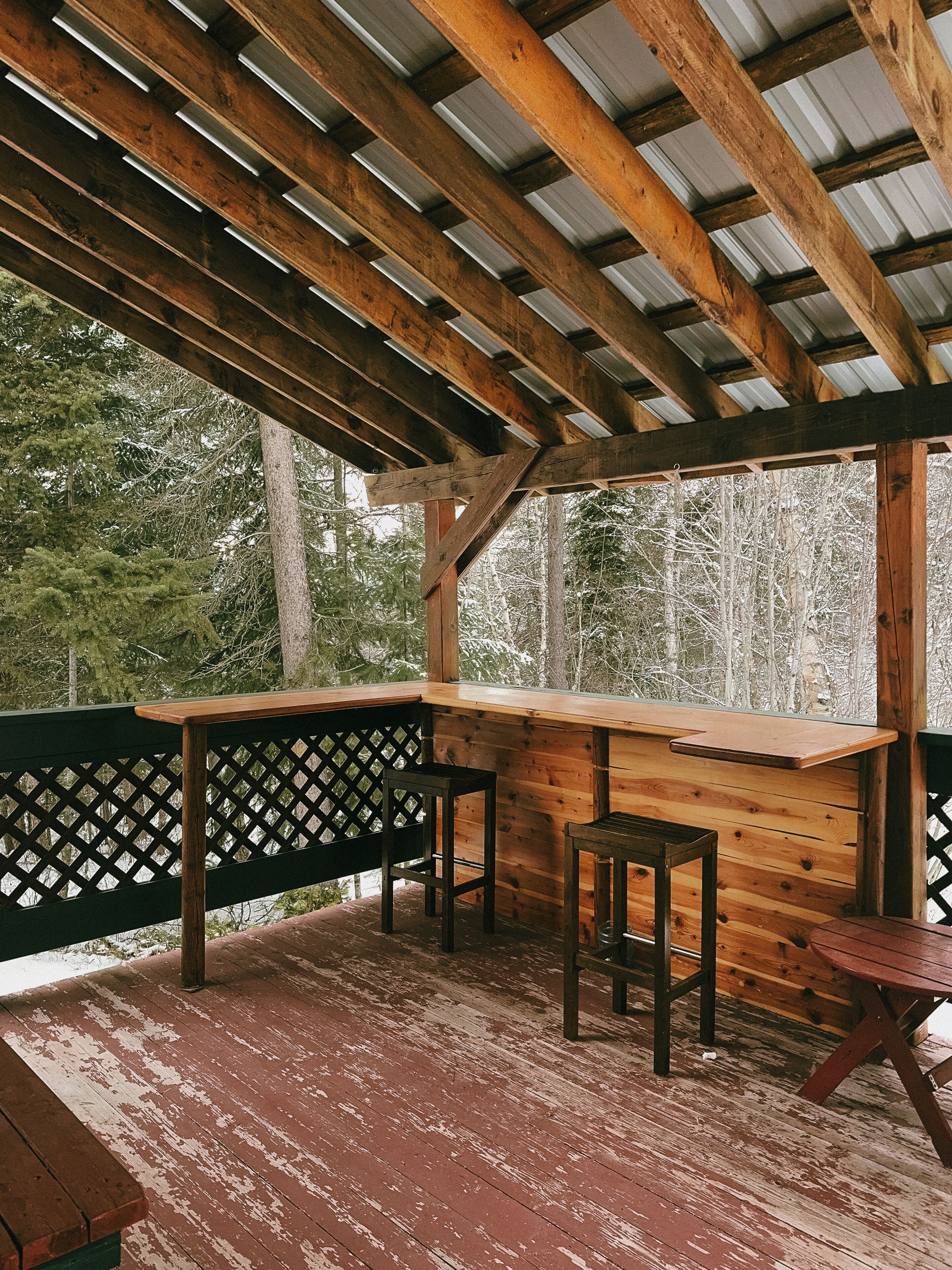 A wooden deck with a bench and a table, surrounded by a metal roof and wooden beams.