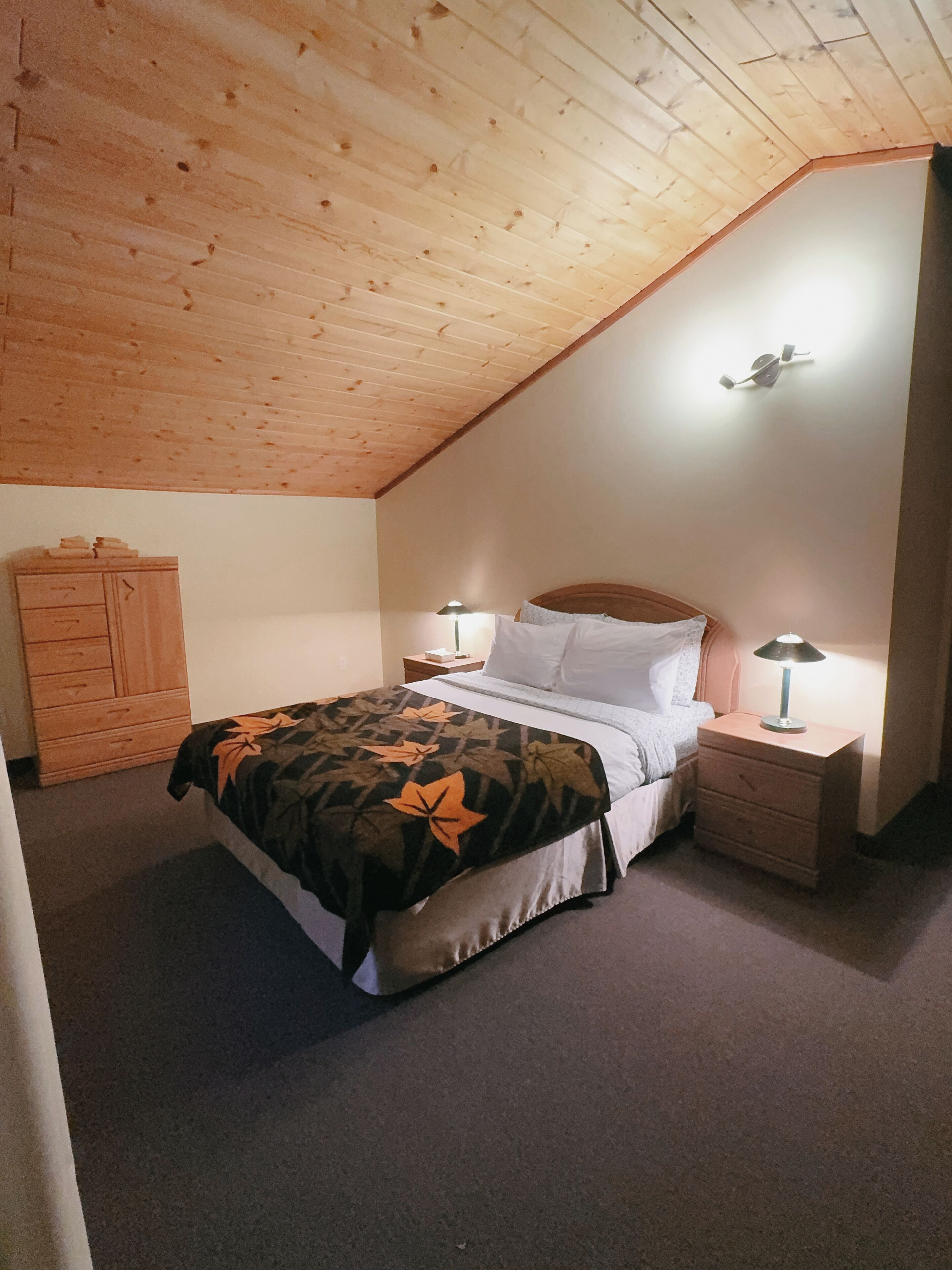 A bedroom with a sloped ceiling, a bed with a patterned bedspread, two bedside tables with lamps, and a wooden wardrobe.