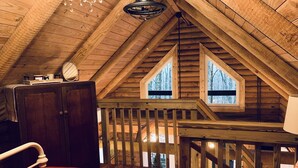 A wooden cabin with a loft bedroom featuring a bed, a dresser, a chandelier, and a view of the forest through two windows.