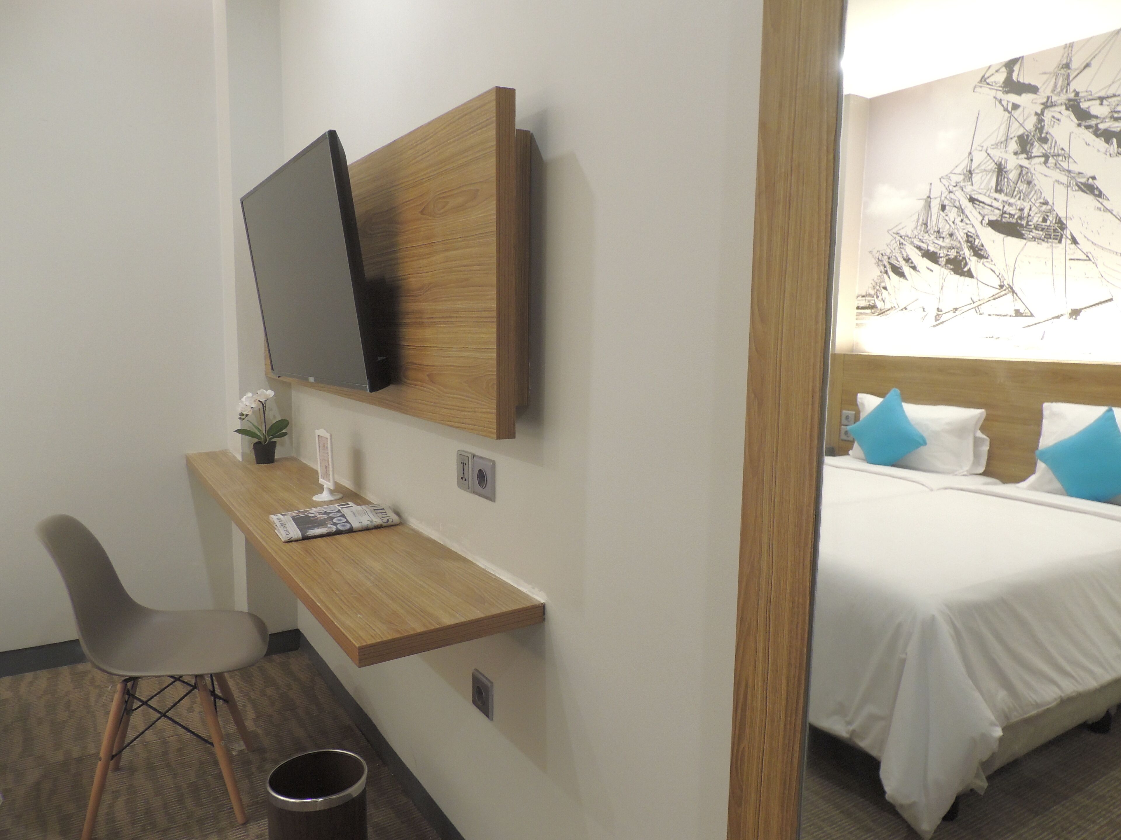 A hotel room with a bed, a desk with a chair, a television mounted on the wall, and a small potted plant.