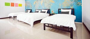 Habitación de hotel con tres camas individuales, cada una con una colcha blanca y almohadas estampadas en blanco y negro. La habitación tiene una pared azul con un mural de motivos vegetales y un suelo blanco.