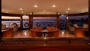 A luxurious cabin with a wooden interior, a central seating area with cushioned chairs, and large windows offering a view of the water. - Jaz Monarch Nile Cruise - Every Monday from Luxor for 07 and 04 Nights - Every Friday From Aswan for 03 Nights (Luxor)