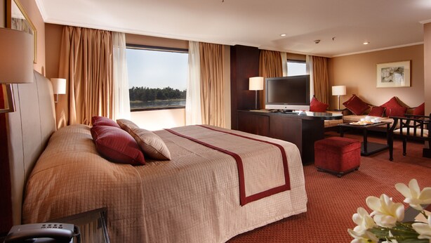 A hotel room with a large bed, a desk with a TV, a sofa, and a view of the outdoors. - Jaz Monarch Nile Cruise - Every Monday from Luxor for 07 and 04 Nights - Every Friday From Aswan for 03 Nights (Luxor)