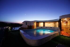 Une piscine à bord incurvé, entourée d’une terrasse en bois et bordée d’un bâtiment à la façade incurvée.