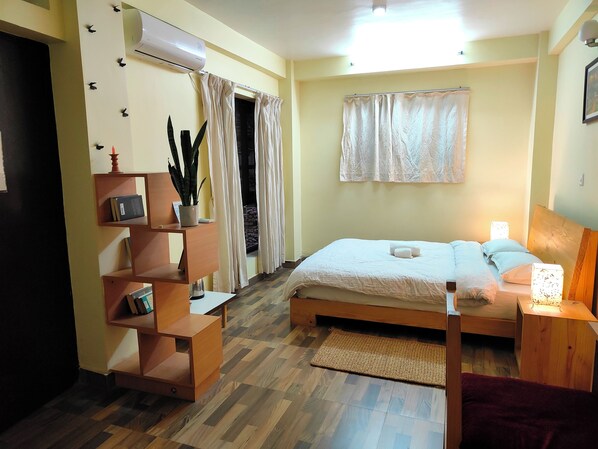 A bedroom with a bed, bedside tables, a wooden shelf unit, a potted plant, and a window with curtains.