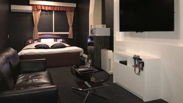 A modern hotel room with a large bed, a flat-screen TV mounted on the wall, a desk with a computer, and a leather sofa. - Hotel Gaudium - Adult Only (Kuki)