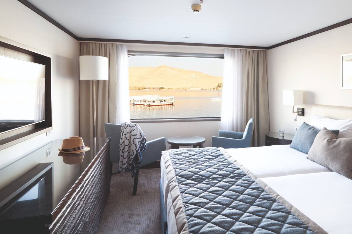 A hotel room with a bed, a desk, a chair, a TV, and a window with a view of a lake and a boat.