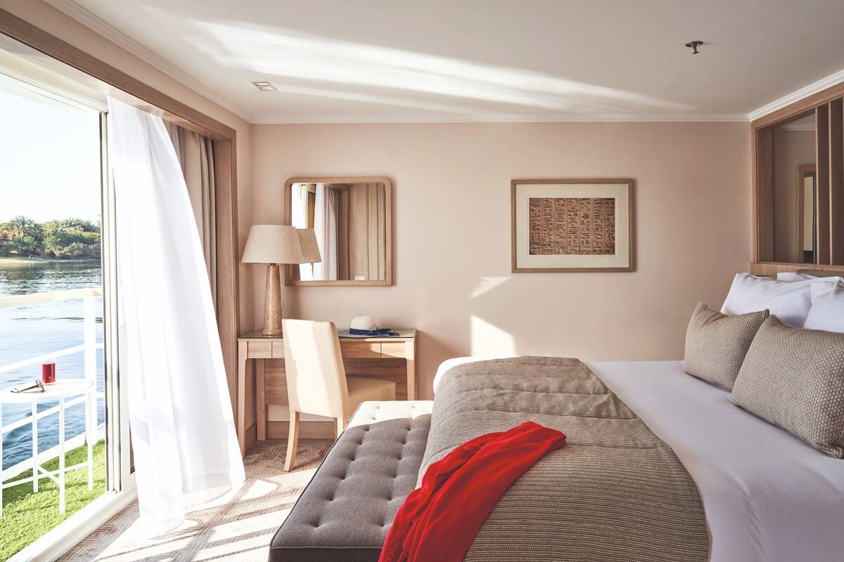 A hotel room with a bed, a desk, a chair, a lamp, and a view of a body of water.