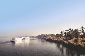 A riverboat on a calm river with a line of palm trees on the right bank. - JAZ Imperial Nile Imperial Cruise - Every Thursday from Luxor- Aswan- Luxor for 07 Nights (Luxor)