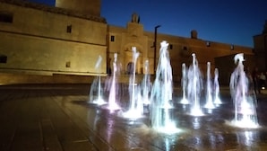A nighttime view of a courtyard with multiple water fountains and a building with a flat roof in the background. - Casa a Corte (Copertino)