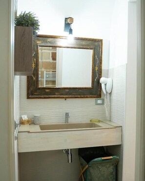 A bathroom with a wooden vanity, a mirror with a decorative frame, a white handheld showerhead, and a trash bin.