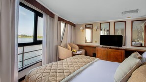 A hotel room with a large bed, a television, a desk, and a view of the water. - Jaz Celebrity Nile Cruise - Every Saturday from Luxor for 07 and 04 Nights - Every Wednesday From Aswan for 03 Nights (Luxor)