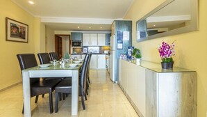 Un comedor moderno con una mesa larga y sillas, una cocina al fondo, y una pequeña planta en maceta sobre una encimera.