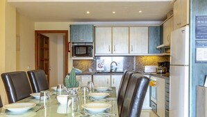 Un comedor con una mesa de vidrio dispuesta para cuatro personas, rodeado de sillas de cuero oscuro. Se ve una cocina con armarios blancos y un microondas al fondo.