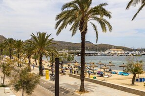 Eine Küstenpromenade mit Palmen, Strandsonnenschirmen und einem Yachthafen im Hintergrund.