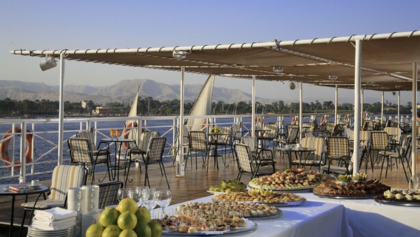 An outdoor dining area with tables and chairs, a buffet with various dishes, and a view of the sea and mountains in the background. - JAZ Crown Emperor Nile Cruise - Every Thursday from Luxor for 07 & 04 Nights - Every Monday from Aswan for 03 Nights (Luxor)