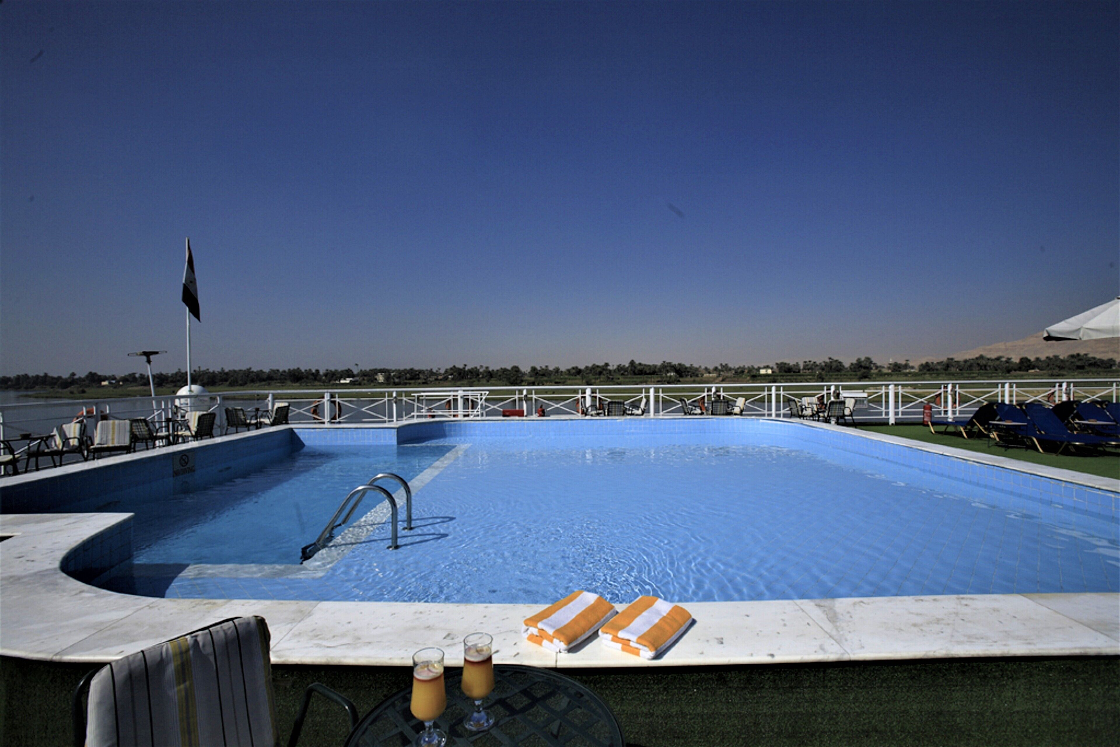 Una piscina con sillones de descanso y sombrillas, una bandera y una mesa con bebidas.