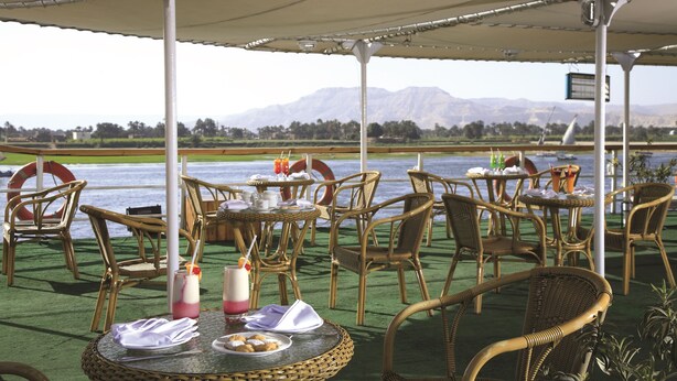 An outdoor dining area with wicker furniture, tables set with plates, glasses, and napkins, overlooking a body of water and mountains. - Jaz Crown Prince Nile Cruise - Every Monday from Luxor for 07 & 04 Nights - Every Friday From Aswan for 03 Nights (Luxor)