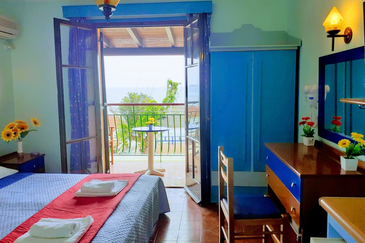 A hotel room with a bed, a desk, a chair, a small table with flowers, and a balcony with a view of the sea.