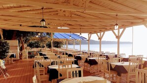 An outdoor dining area with white tables and chairs, a wooden deck, and a view of the water. - Medusa Studios (Kefalonia)
