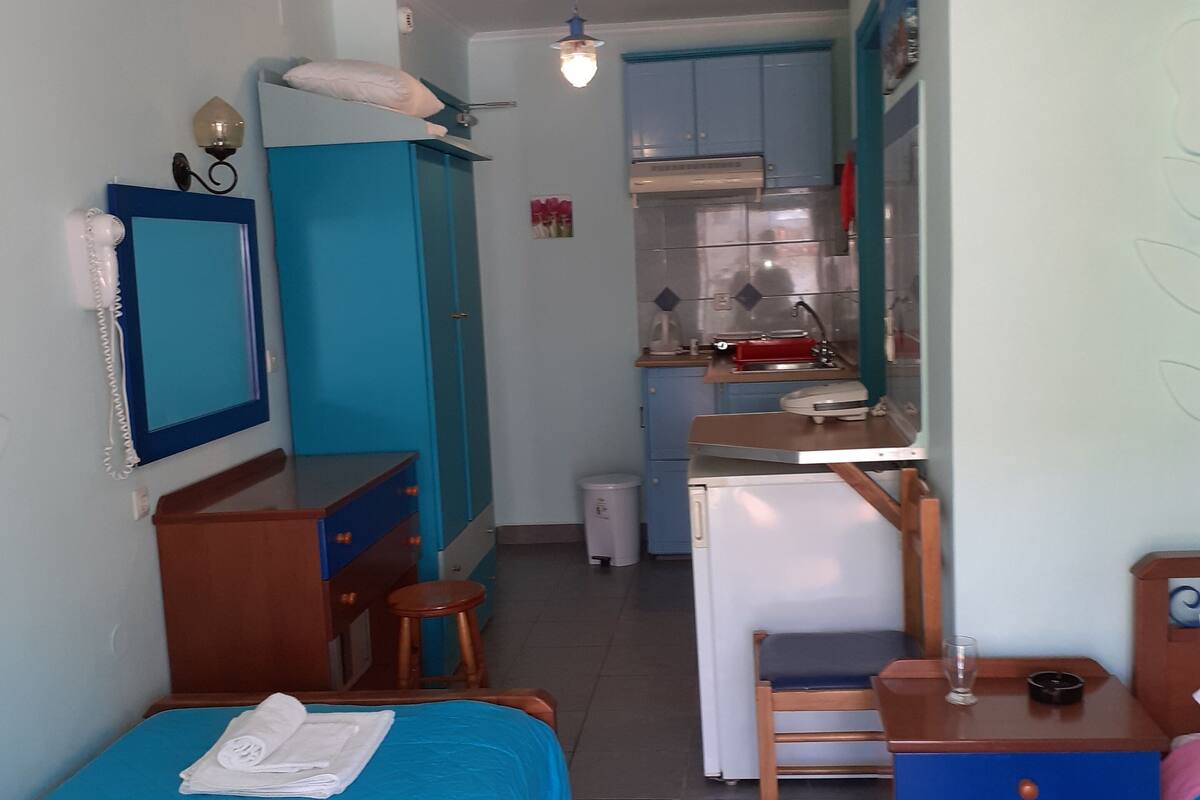 A compact hotel room with a single bed, a desk with a chair, a wardrobe, a small kitchenette, and a bathroom.