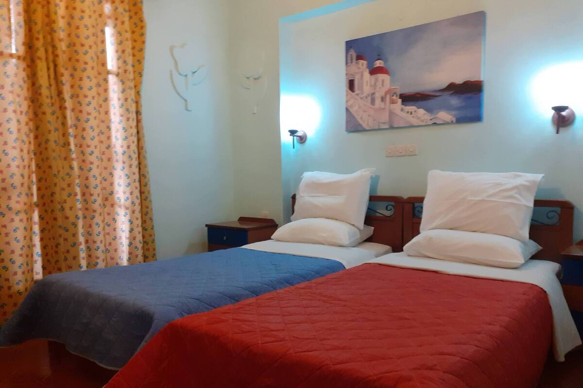 A hotel room with two beds, each with a different colored bedspread, a painting on the wall, and a curtain with a floral pattern.